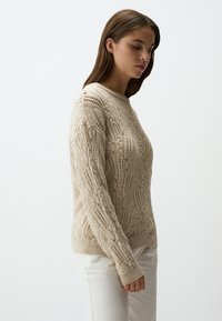 Beige gestrickter Pullover mit einem strukturierten Zopfmuster. Hat einen runden Halsausschnitt und gerippte Bündchen, kombiniert mit weißen Hosen.