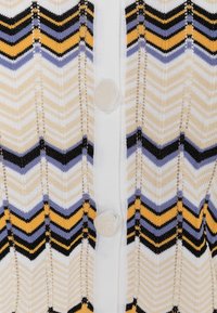 Cárdigan de punto con un patrón de chevron en crema, negro, amarillo y lavanda. Presenta tres grandes botones de crema en la parte frontal. Textura suave.