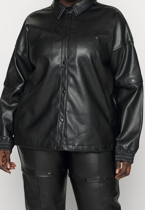 Person trägt schwarze Lederjacke mit Knopfleiste vorne und passende schwarze Lederhose mit Druckknopfdetails an den Oberschenkeln.