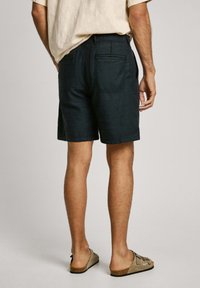 Pantalones cortos de color verde oscuro, confeccionados en una tela ligera, con un diseño de frente plano, bolsillos traseros y un corte recto. Llevados con sandalias de color claro.
