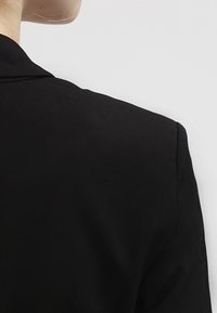 Czarny żakiet o dopasowanym kroju, gładkiej fakturze i wyraźnie zarysowanych ramionach. Materiał wydaje się lekki i elegancki.