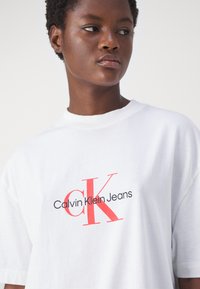 Bílý bavlněný tričko s červeným a černým grafickým potiskem "Calvin Klein Jeans" a velkým logem "CK" na přední straně.