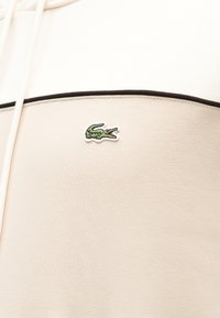 Μπεζ ένδυμα με μαύρο περίγραμμα που διαθέτει ένα μικρό πράσινο κεντημένο λογότυπο κροκόδειλου στο ύψος του στήθους.