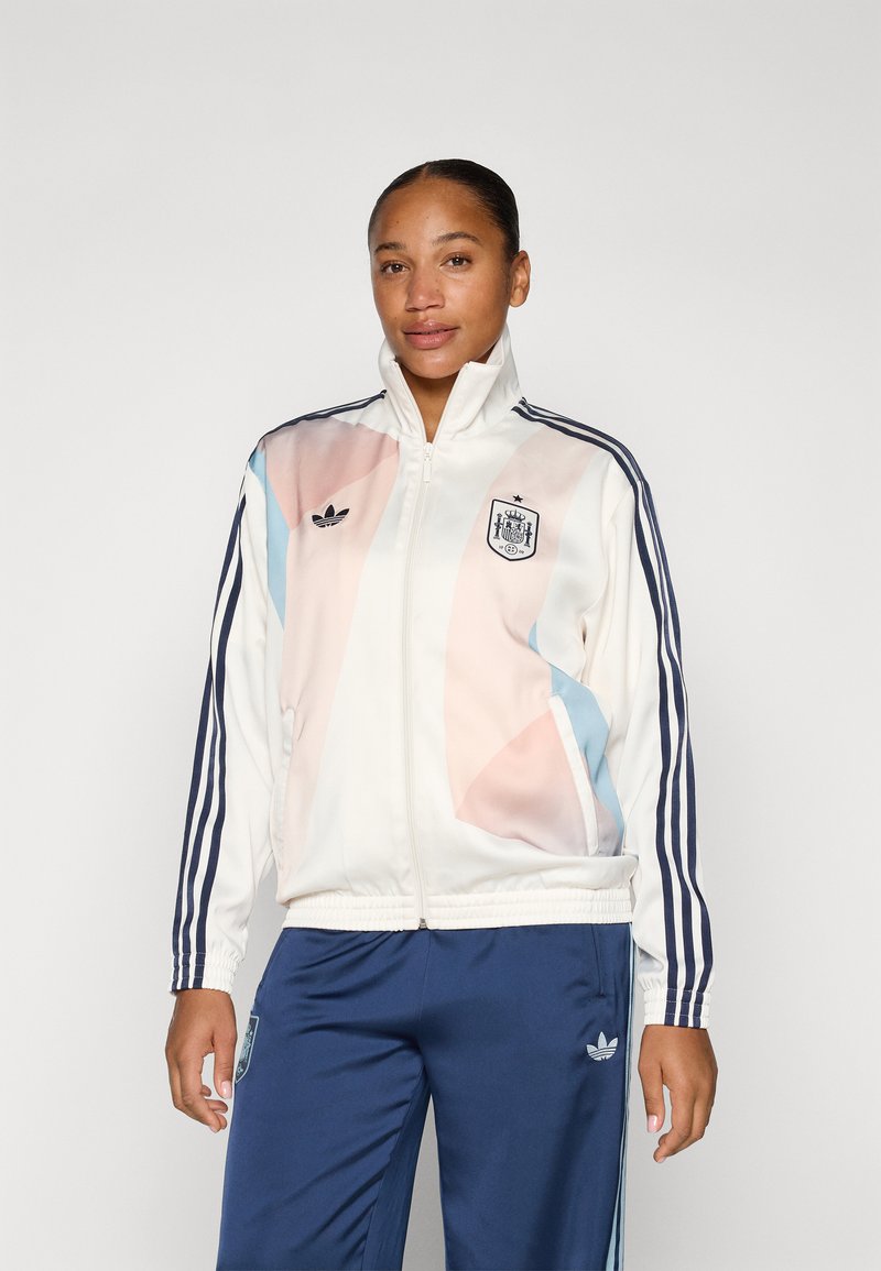 Bílá bunda s růžovými a modrými geometrickými vzory, s vysokým límcem, zipovým zapínáním a logem Adidas na hrudi a rukávech.
