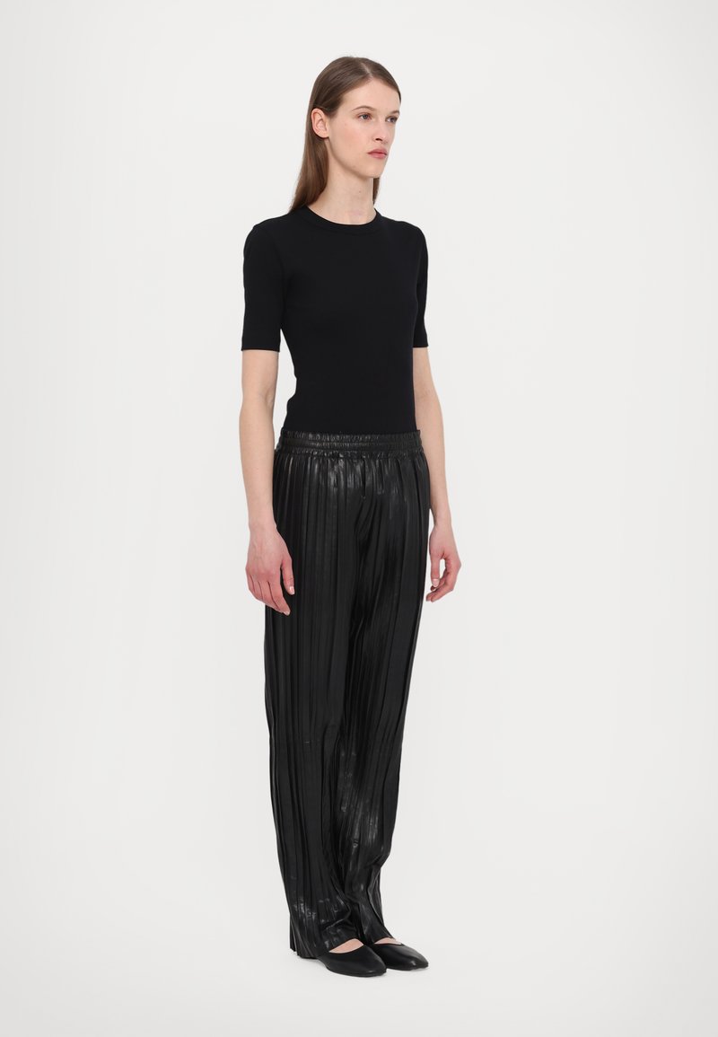 Svart åtsittande topp med kort ärm kombinerat med glänsande svarta plisserade byxor med vida ben, som har ett elastiskt midjeband och slät yta. Svarta ballerina-skor.