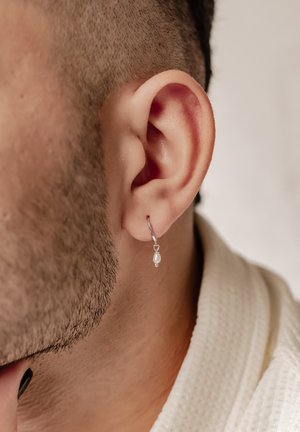 Boucle d'oreille en argent avec une petite perle blanche pendante. Vue en gros plan de l'oreille et de la peau texturée, mettant en avant les détails de la boucle.