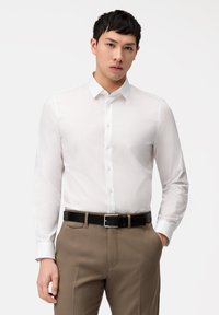 Weißes Langarmhemd mit Button-Down-Kragen, glatter Textur und tailliertem Design, kombiniert mit beigen Hosen und einem schwarzen Gürtel.