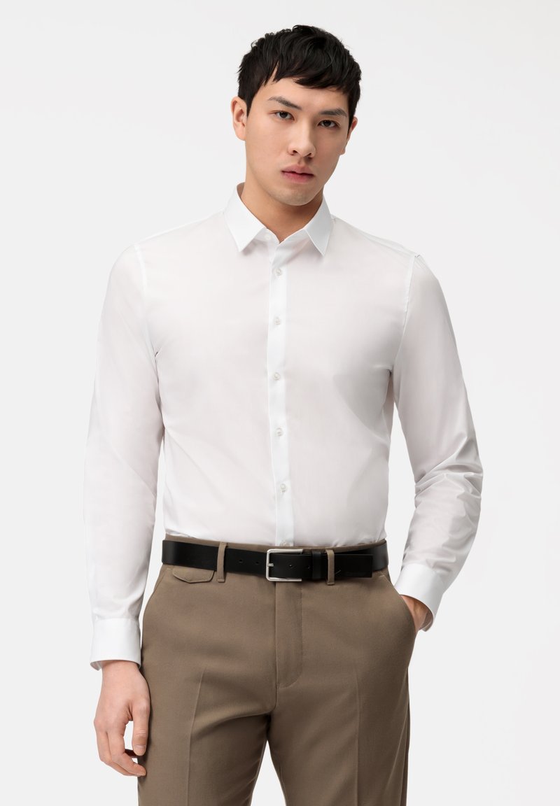 Weißes Langarmhemd mit Button-Down-Kragen, glatter Textur und tailliertem Design, kombiniert mit beigen Hosen und einem schwarzen Gürtel.