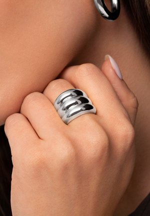 Anillo de plata con tres bandas semicirculares pulidas que exhiben un acabado suave y reflectante. Mano apoyada contra un fondo neutral.