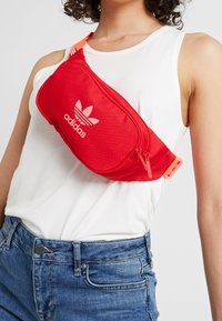 Červená nylonová ledvinka s přední kapsou na zip, s bílým logem Adidas a nastavitelným popruhem. Texturovaný materiál s elegantním designem.