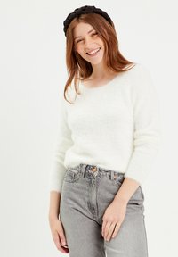 Pull blanc en laine duveteux avec un décolleté large, manches longues, associé à un jean gris taille haute. Le modèle porte un serre-tête noir tressé.