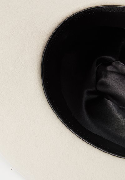 Chapéu de feltro creme com um forro interior de cetim preto e uma faixa preta costurada à volta da aba, apresentando uma textura lisa e forma arredondada.