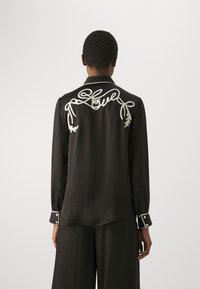 Camicia di seta nera con dettagli ricamati bianchi sul retro, che presentano foglie e anelli, e polsini con bottoni a contrasto.
