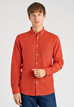 Czerwona koszula na guziki z długimi rękawami, pojedynczą kieszenią na piersi i kołnierzykiem. Tkanina wydaje się mieć teksturę i lekki połysk. Do tego ciemnoniebieskie dżinsy.
