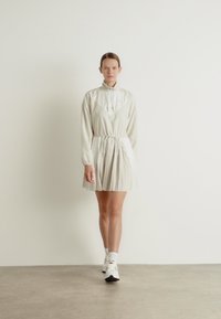 Ljusbeige klänning med långa ärmar, plisserad kjol och insvängd midja. Har en hög krage och en kontrasterande triangel-bystdesign.