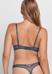 Woman with long blonde hair seen from behind wearing teal mesh bra and matching thong underwear against a plain light background.