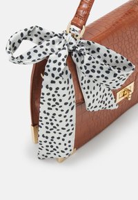 Hnědá kožená kabelka s texturovaným krokodýlím vzorem, zdobená bílým šátkem s černými puntíky přivázaným k rukojeti a zlatými kovovými doplňky.