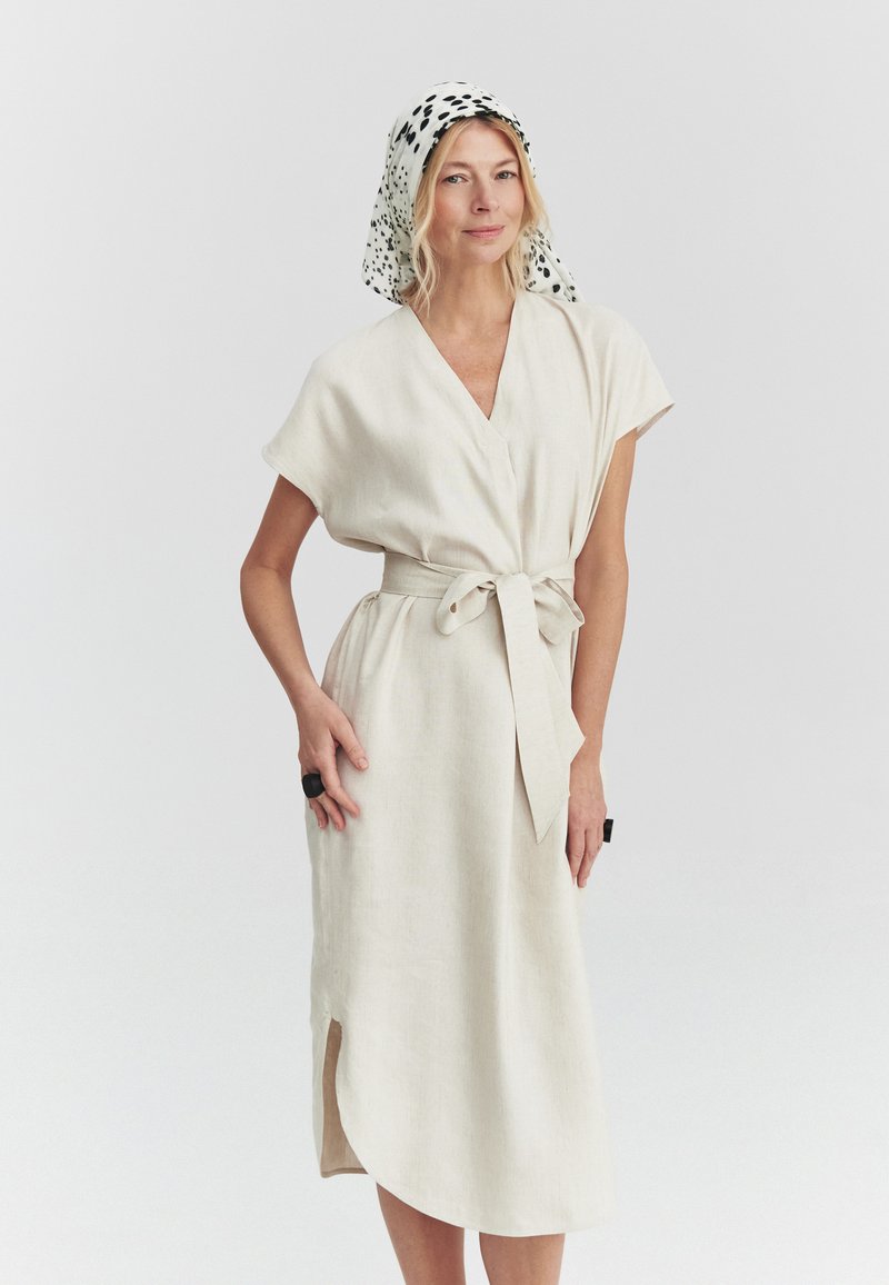 Femme portant une robe portefeuille beige clair avec une ceinture nouée à la taille et un foulard blanc à pois noirs, debout devant un fond uni.