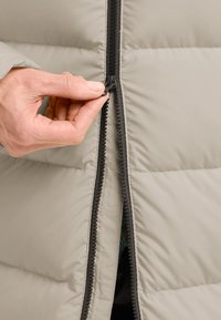 Lichtgrijze gewatteerde jas met een zwarte rits. Een hand grijpt de ritsen van de rits, wat een kleurrijke binnenvoering onthult.