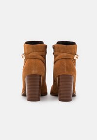Botas de tobillo de ante marrón con tacón apilado. Presentan hebillas en tono dorado y cremalleras laterales, textura suave y punta redondeada.