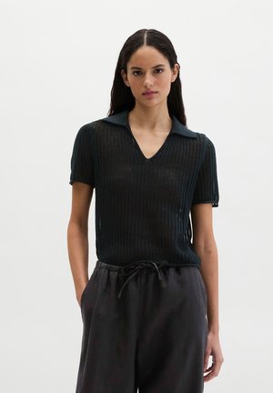 Kobieta o długich ciemnych włosach ubrana w czarną dzianinową bluzkę z krótkim rękawem i kołnierzykiem oraz czarne spodnie ze sznurkiem, stoi z jedną ręką w kieszeni.