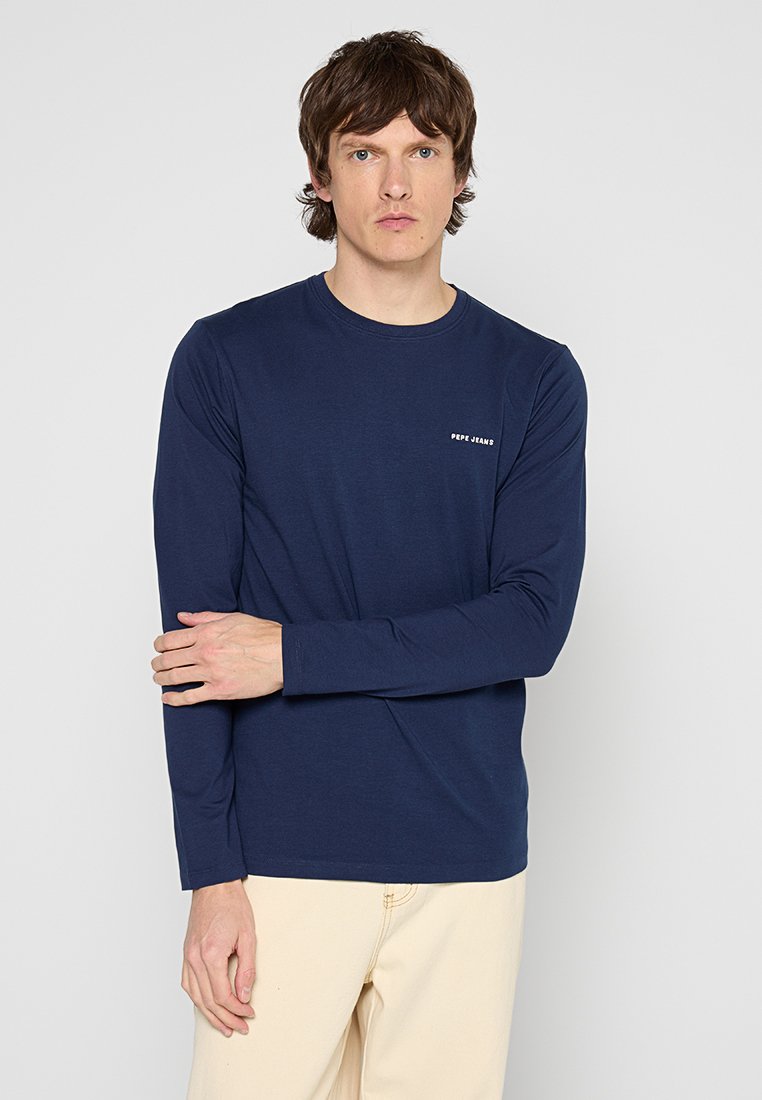 Camiseta de manga larga azul marino hecha de tejido suave, con escote en forma de círculo y pequeño logo blanco en el lado izquierdo del pecho. Llevada con pantalones de color claro.