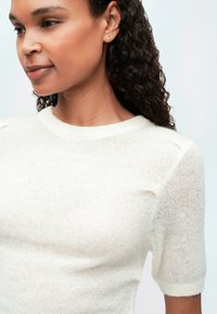 Γυναίκα με σγουρά σκούρα μαλλιά, φορώντας λευκό πλεκτό μπλουζάκι κοντού μανικιού, κοιτώντας ελαφρώς στο πλάι σε μία απλή φωτεινή φόντο.