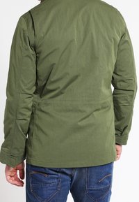 Grön, lätt vikjacka med en slät textur, som har långa ärmar och en enkel, rak skärning. Bakifrån syns en ren design.