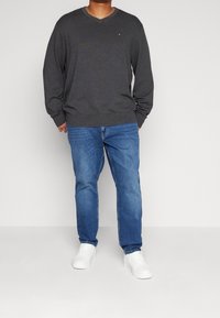 Grauer V-Ausschnitt-Pullover mit langen Ärmeln, kombiniert mit blauen Jeans und weißen Sneakers. Der Pullover zeigt ein dezentes Branding auf der Brust.
