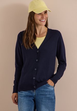 Femme souriante portant une casquette jaune, un top jaune, un cardigan bleu marine boutonné et un jean bleu, une main dans la poche sur fond beige.