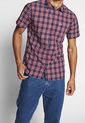 Mann trägt ein kurzärmeliges, rot-weiß-navy kariertes Hemd mit Knöpfen und blaue Jeans, steht vor einem weißen Hintergrund.