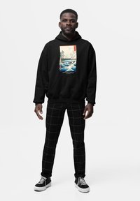 Felpa nera con grafica frontale raffigurante una scena paesaggistica. Abbinata a pantaloni neri a scacchi e sneakers nere con suole bianche.
