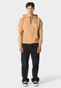 Sudadera marrón clara con cuerda, bolsillo frontal y logo en el pecho. Combinada con pantalones negros y zapatos negros con suelas blancas.