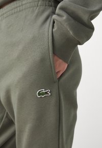 Olivově zelené tepláky s hladkou texturou, na levé kapsě vyšívané logo krokodýla a elastické manžety na kotnících.