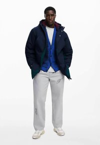 Mörkblå vadderad jacka med huva, blå texturerad väst, vit skjorta, grå byxor med tryckt detalj och beige sneakers. Modern, avslappnad lager-på-lager-stil.