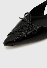 Black patent leather shoe with a pointed toe, featuring decorative laces and a cut-out back design. Smooth texture with a glossy finish.