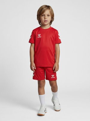Ung dreng står iført et rødt Hummel sportstøj med hvide sokker og hvide sneakers på en ensfarvet lys baggrund.