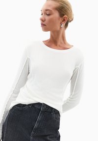 Biała koszula z długim rękawem, wykonana z miękkiej tkaniny, z okrągłym dekoltem, zestawiona z ciemnoniebieskimi dżinsami. Prosty krój, gładka tekstura.