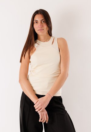 Donna con capelli castani lunghi che indossa una canotta beige e pantaloni neri, in piedi con le mani intrecciate davanti a uno sfondo bianco.