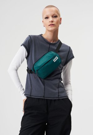 Person mit kurzen Haaren, der übereinander getragene Lang- und Kurzarmshirts, schwarze Hosen und eine türkisfarbene Umhängetasche mit der Aufschrift "A-HoR" trägt.