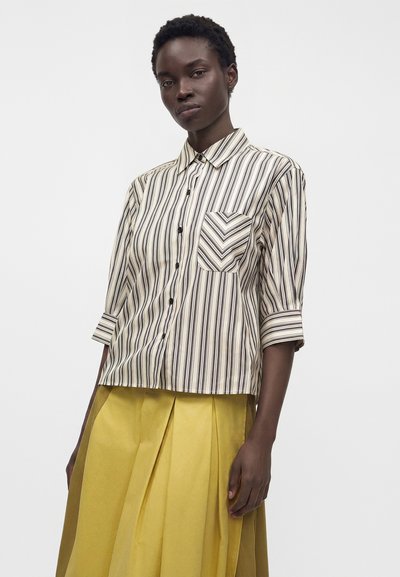 Femme portant une chemise boutonnée beige et noire à rayures verticales avec des manches trois-quarts et une jupe plissée jaune.
