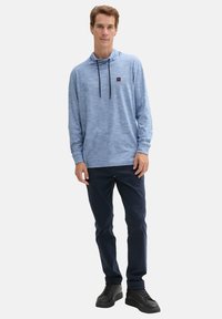 Felpa cappuccio blu chiaro melangiato con maniche lunghe, cordini neri regolabili e una piccola patch con logo. Indossata con pantaloni blu navy e sneakers nere.
