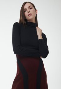 Zwarte coltrui gecombineerd met een bordeauxrode jurk met zwarte zijaccenten. Gladde textuur, aangesloten vorm en lange mouwen.