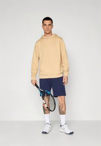 Bēšas krāsas džemperis ar kapuci, tumši zili šorti, balti zeķes un sporta apavi. Tur rokās tenisa raketi ar ziliem akcentiem. Redzamas tetovējumu zīmes uz kājām.