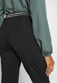 Svarta byxor med en elegant design har en glänsande silverdetalj vid midjan. Tyget ser slätt ut, kombinerat med en texturerad grön blus.