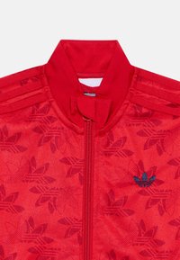 Sarkans zip-up džemperis ar ziedu reljefu, augstu apkakli un tumši zilā Adidas logo uz krūtīm. Struktūras audums un sportiskais dizains.