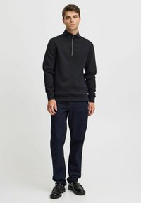 Sweat-shirt noir zippé avec un col haut, des manches longues et une texture, associé à un jean en denim foncé et des chaussures noires.