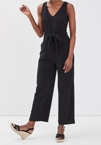 Combinaison noire avec un décolleté en V, des jambes larges et une ceinture nouée à la taille. Portée avec des sandales espadrilles noires à bride de cheville.