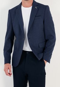 Marinblå blazer i strukturerad tyg med en enkel knapp, slagkrage och bröstficka. Bärs över en vit skjorta.