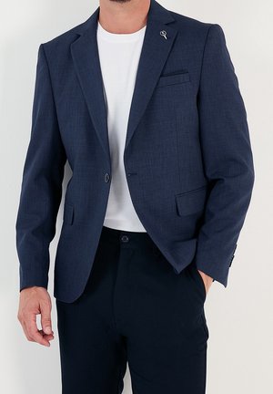 Blazer azul marino de tejido texturizado, con un solo botón, solapas de muesca y un bolsillo en el pecho. Llevado sobre una camisa blanca.